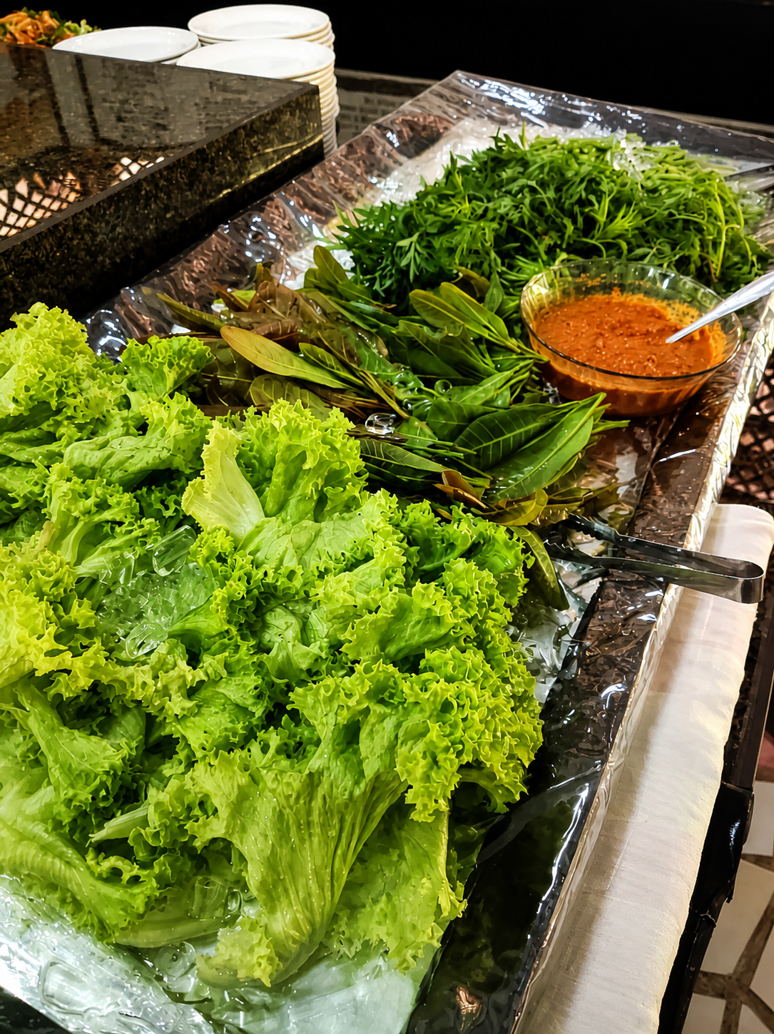 A tray of curly lettuce and fresh herbs laid on crushed ice beside a glass bowl of sambal with a spoon in it.
