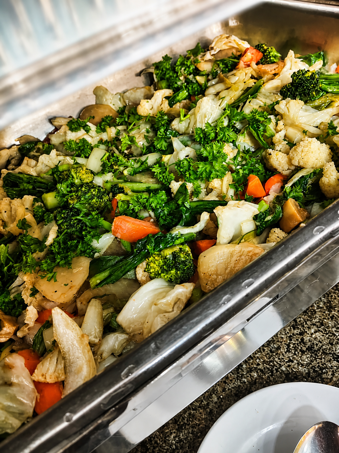 Mixed cooked vegetables — cauliflower, broccoli, carrot and cabbage — under chopped parsley in a metal serving tray.