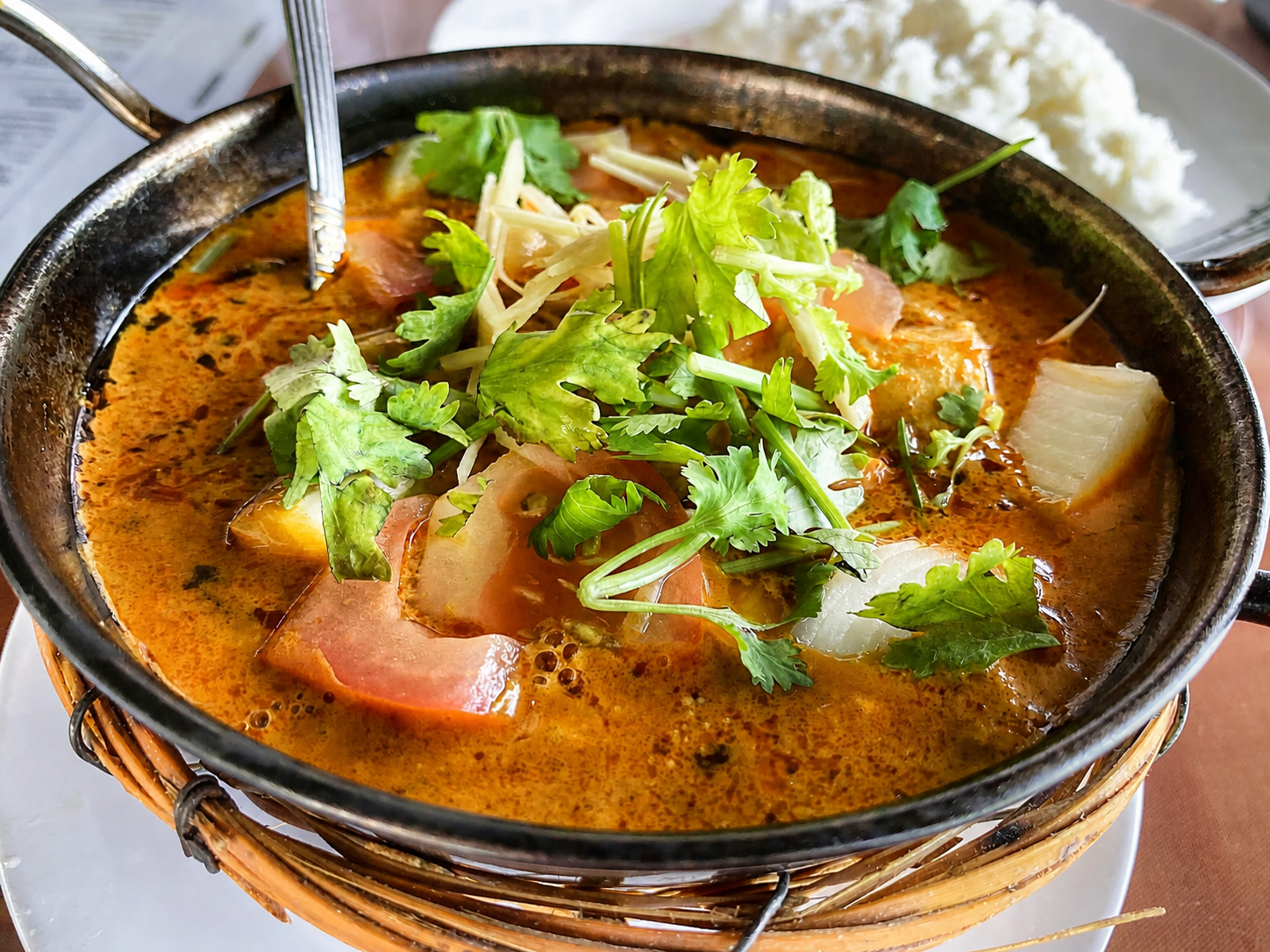 A masala in a black karahi topped with coriander and tomato, a bowl of steamed rice beside it.