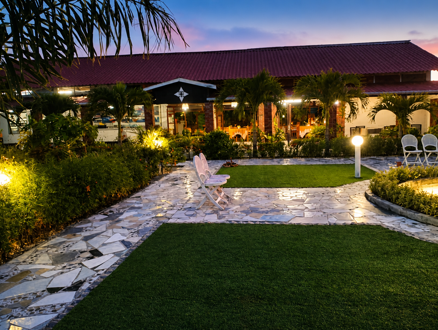 The lit building at nightfall seen across the lawn, with a white bench on the paved path and palms along the front of the veranda.