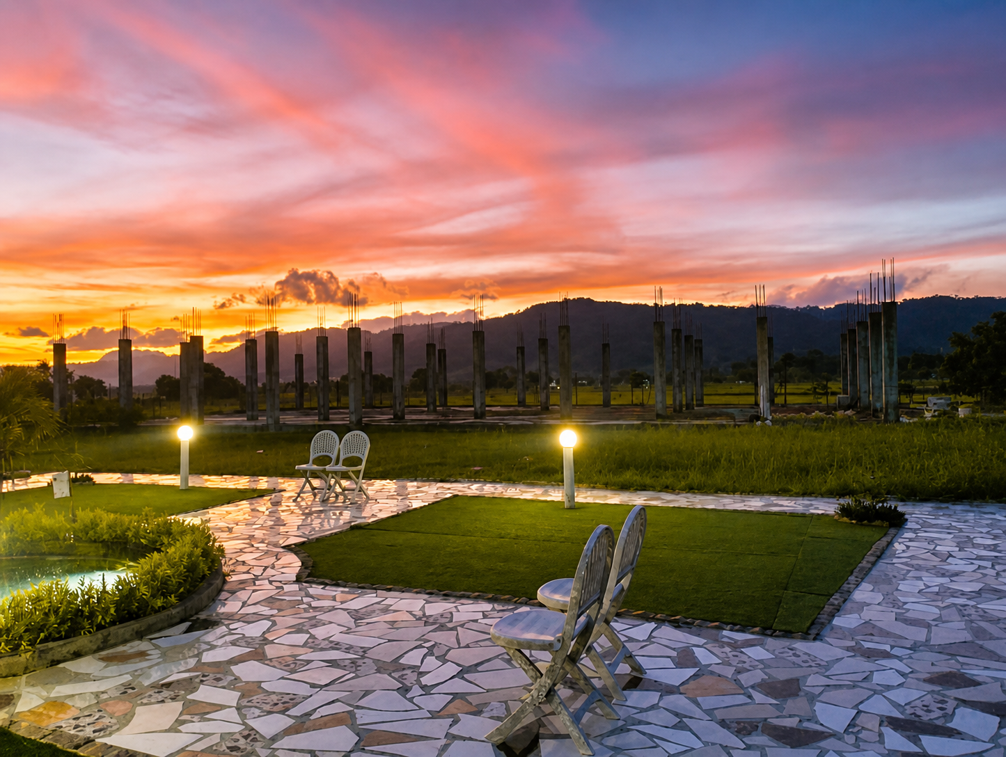Dusk over the garden terrace: white chairs on crazy-paved stone beside clipped lawn, under a pink and orange sky with hills on the horizon.