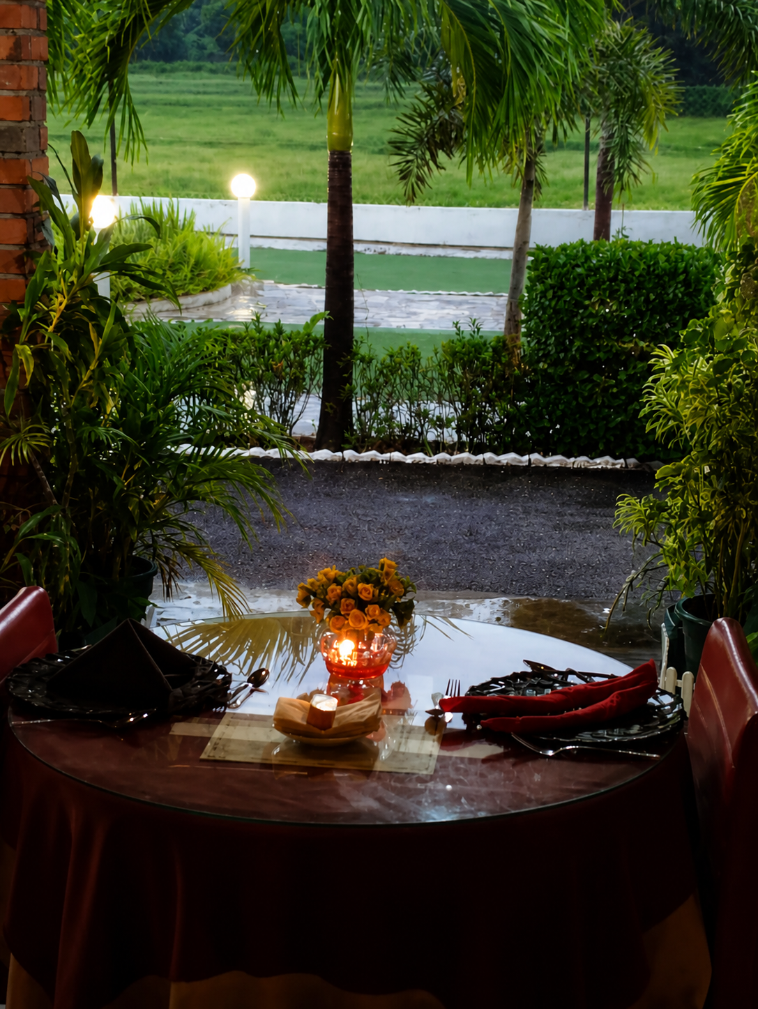 A laid round table at the open edge of the dining room at dusk, looking out past palms and a clipped hedge to the lawn and the field beyond.