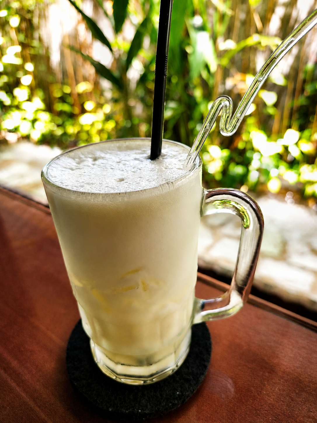 A tall glass mug of pale, frothy lassi with a straw, standing on a dark coaster on a polished table edge with bright greenery behind it.