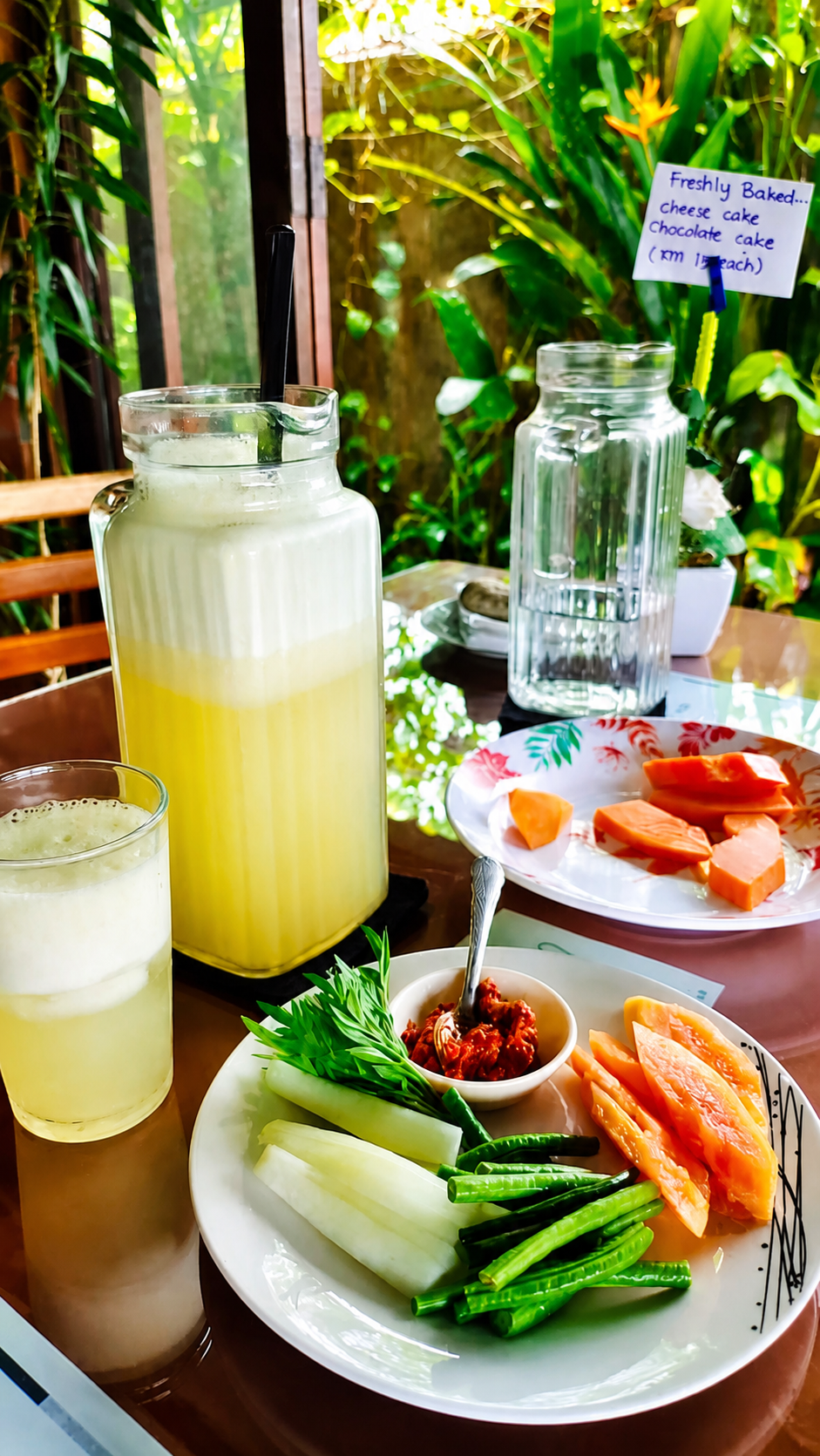 A jug and a tall glass of cloudy lime juice on a garden table, beside a plate of cut raw vegetables and a small dish of sambal.