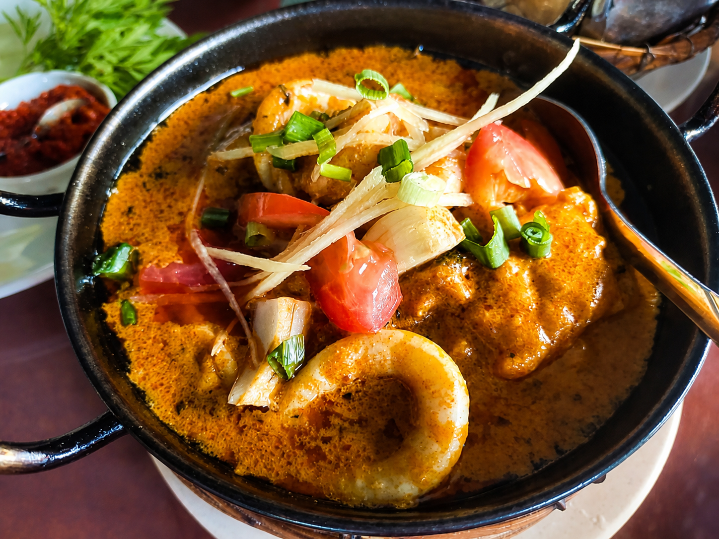 A golden seafood curry with a ring of squid, tomato and spring onion in a black karahi.