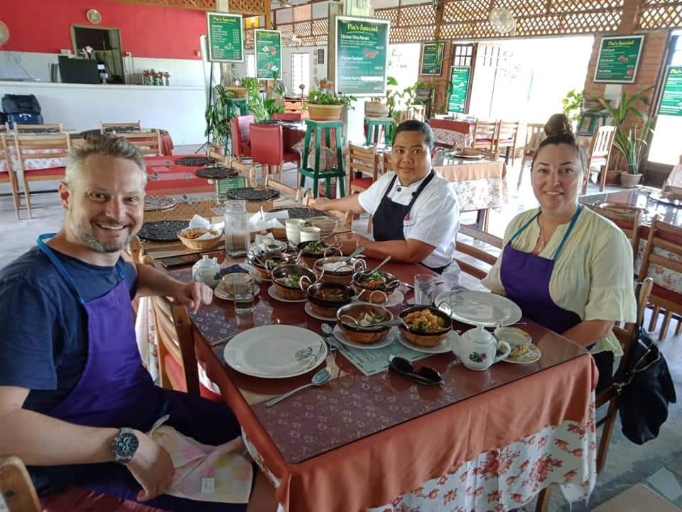 The chef standing behind a table of a dozen finished dishes with two class participants in aprons seated on either side.