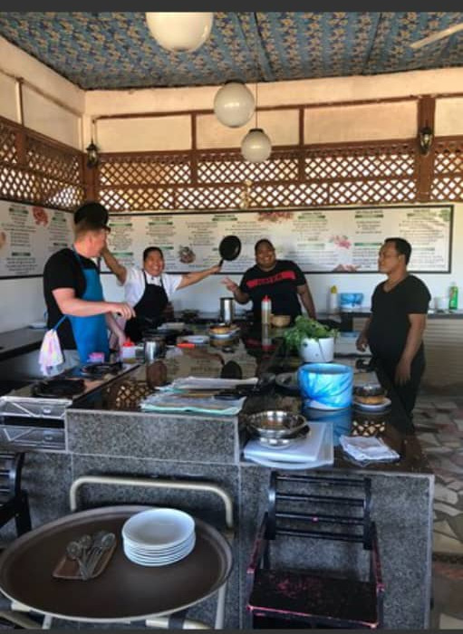A participant in an apron at the open preparation counter while the chef lifts a frying pan beside him.