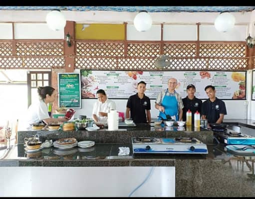 A class group lined up along the open preparation counter with the kitchen team, a gas burner and pans in front of them.