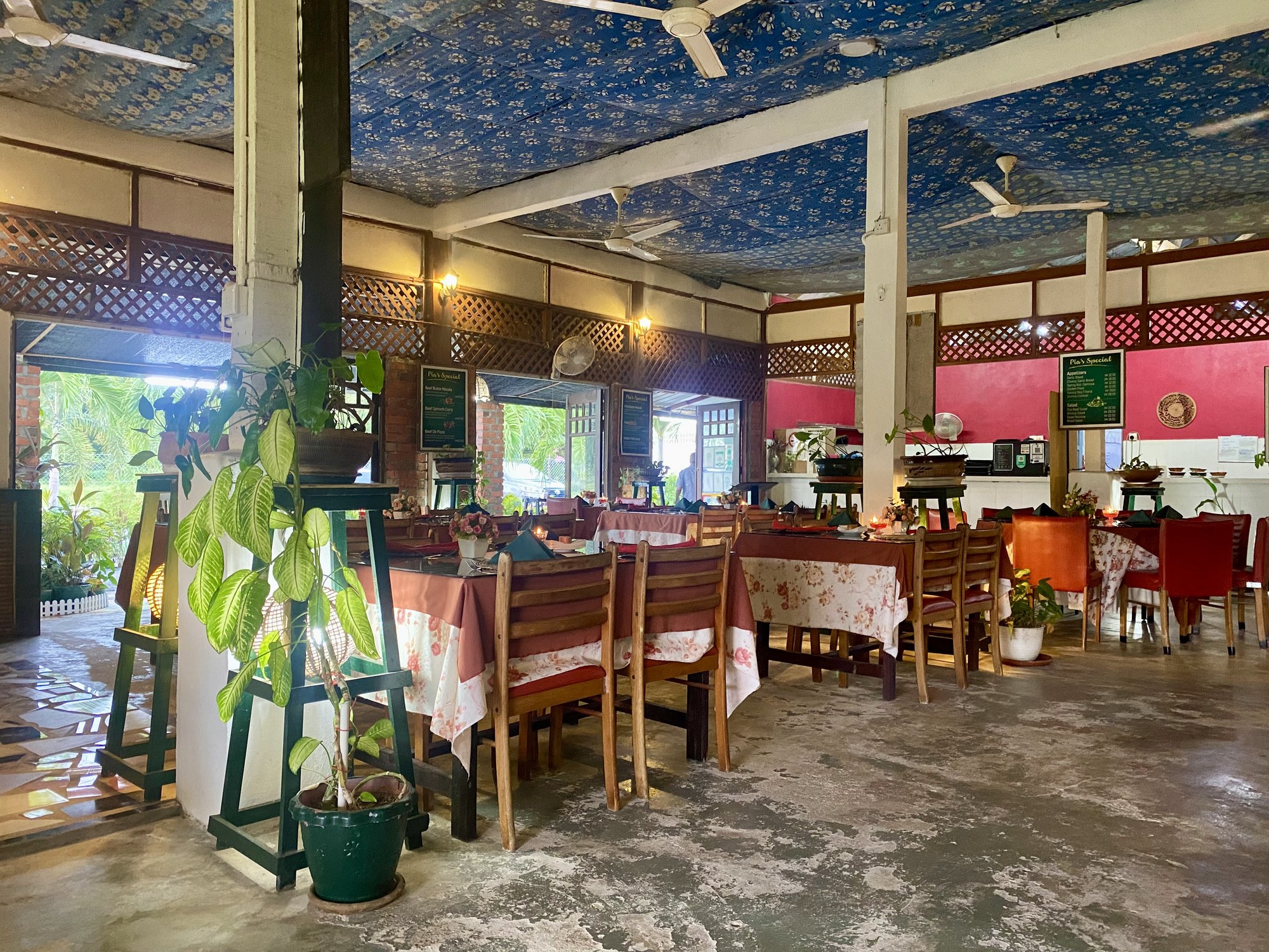 The open dining room at Pia's the Padi: long tables under a printed ceiling cloth, wooden lattice screens, and greenery through the open sides.
