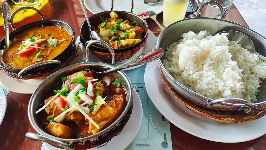 Four karahi pans of different curries set around a bowl of rice on a laid table.
