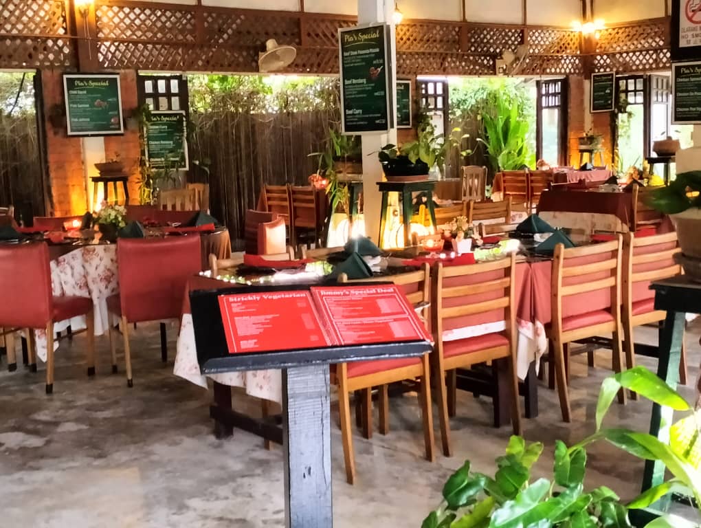 The open dining room by day: long tables under a lattice ceiling, wooden chairs, potted greenery and a bamboo screen along the far side.