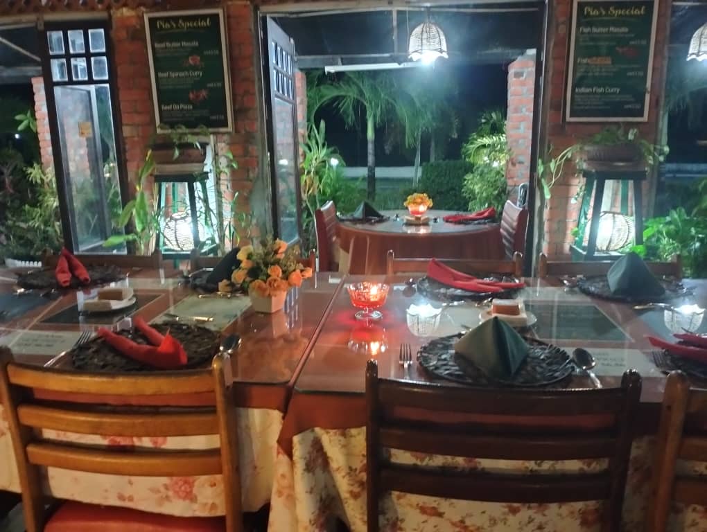 Tables laid with candles, flowers and folded napkins in the evening, open on both sides to a lit garden of palms and brick columns.