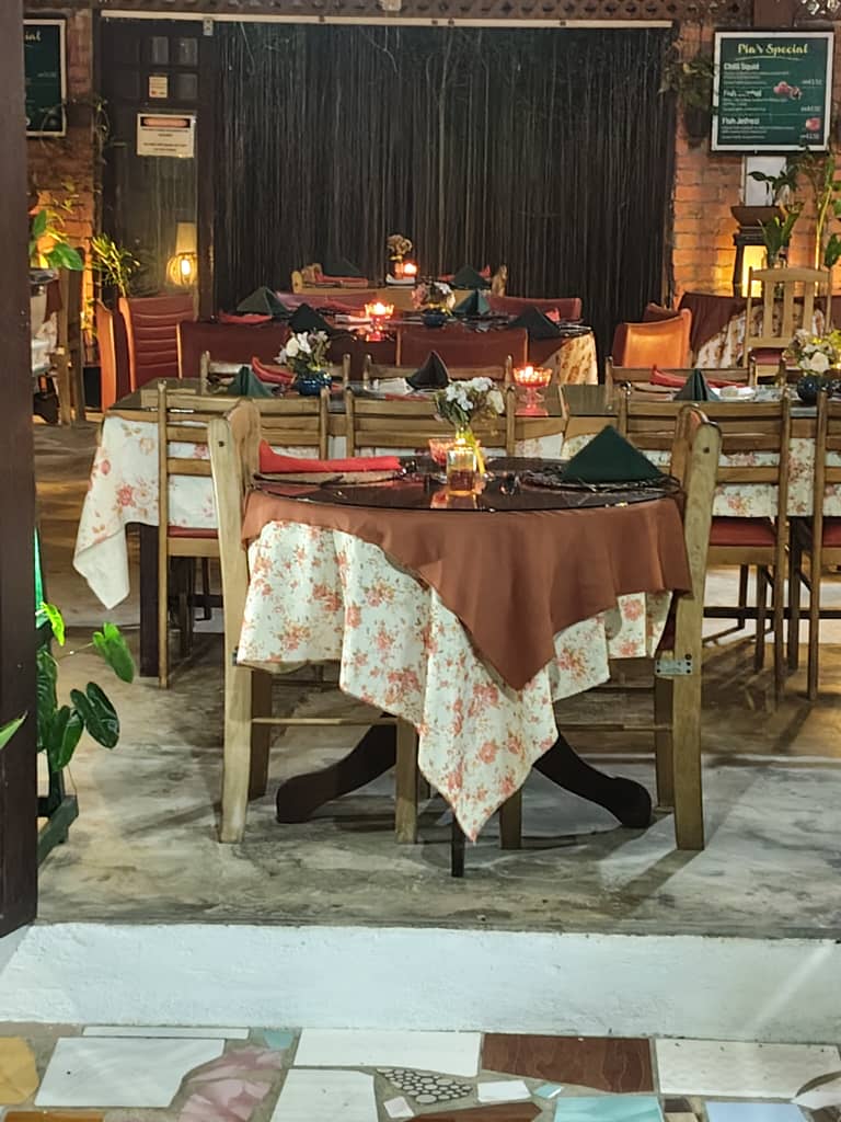 Round and square tables laid with floral cloths, green napkins and candles at dusk, standing on the mosaic-tiled floor at the edge of the room.