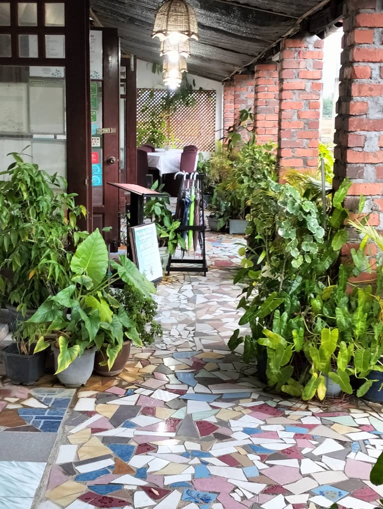 A covered walkway paved in coloured mosaic tiles, lined with potted ferns and leafy plants between brick columns, with a rattan lamp overhead.