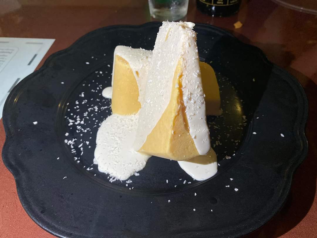 Two triangular slices of frozen mango kulfi dusted with coconut on a dark scalloped plate.
