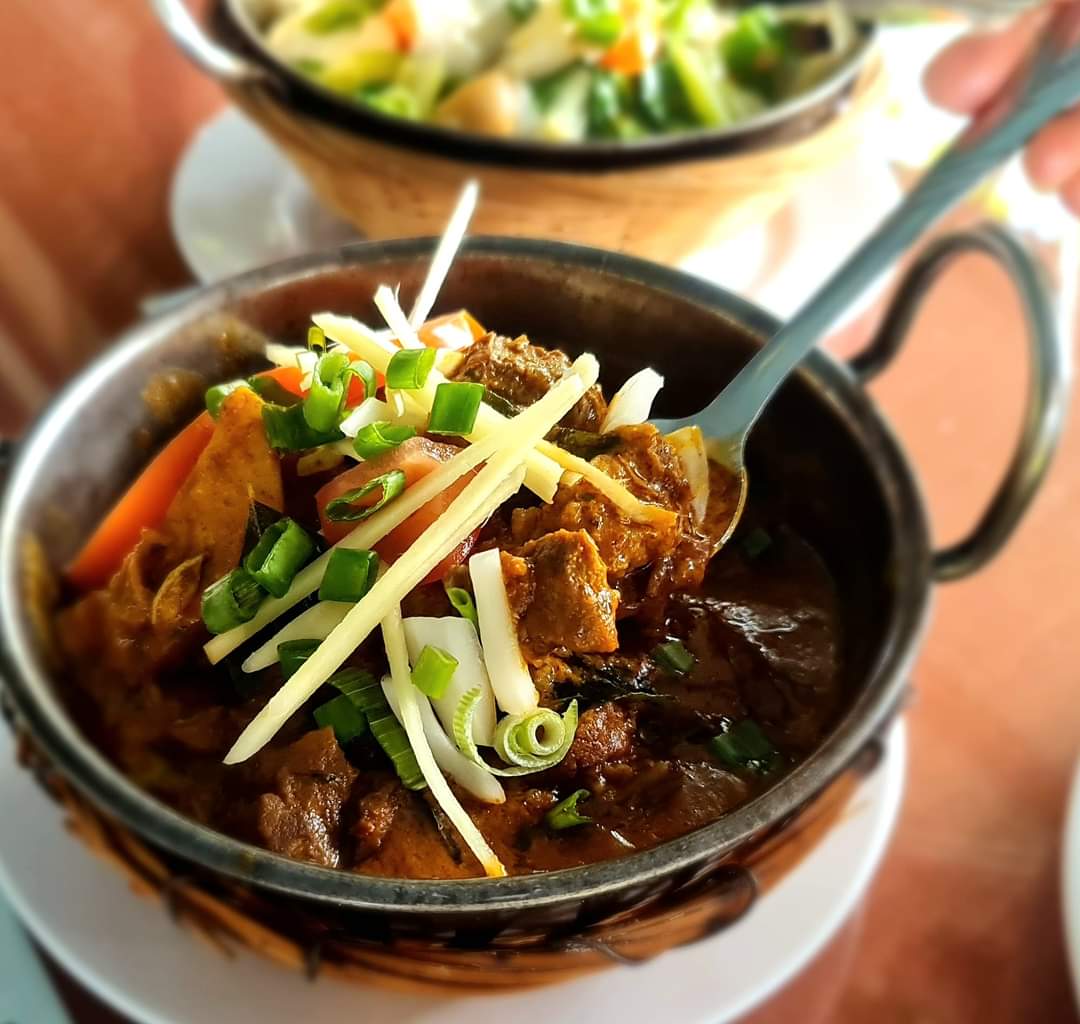 A dark, slow-cooked meat curry in a karahi, finished with ginger and spring onion, a bowl of salad behind it.